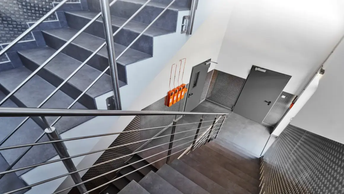 Moderner Treppenaufgang mit Metallgeländern und grauen Stufen. An der Wand hängen orangefarbene Geräte, die Teil des Rauchabzug Treppenhaus Systems sind.
