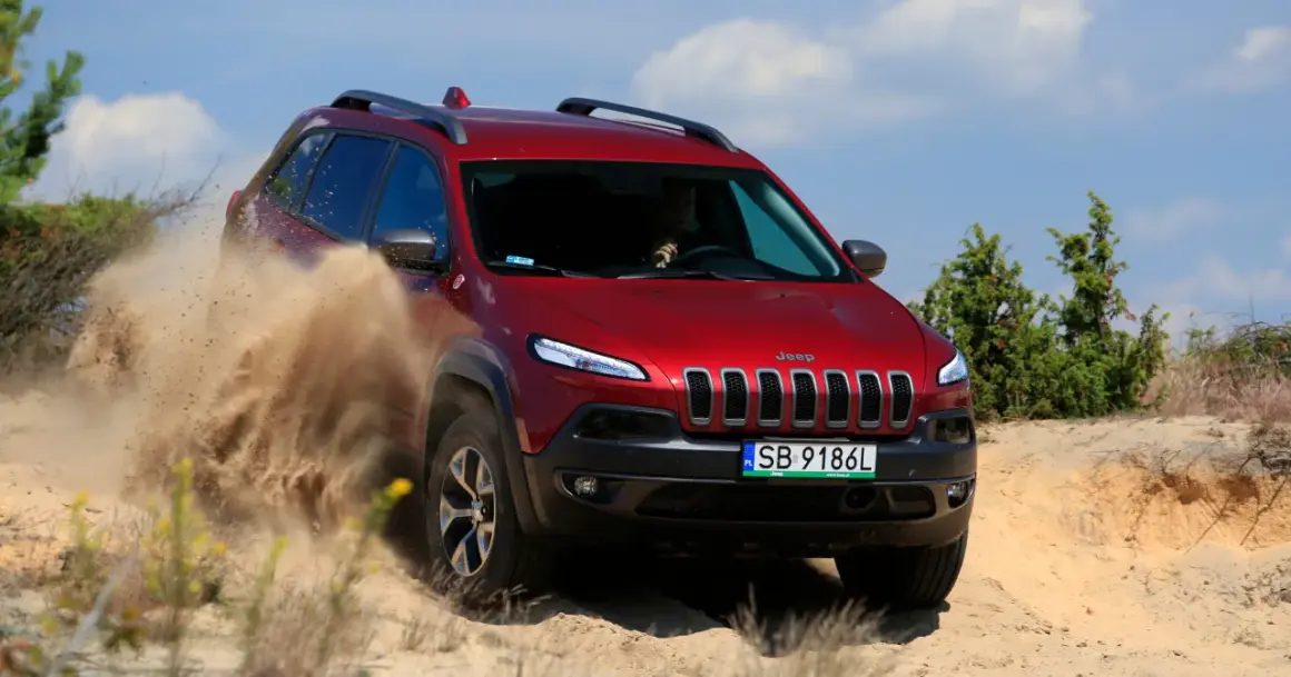 Czerwony Jeep Cherokee Trailhawk z impetem przemierza piaszczysty teren, wzbijając tumany piasku.