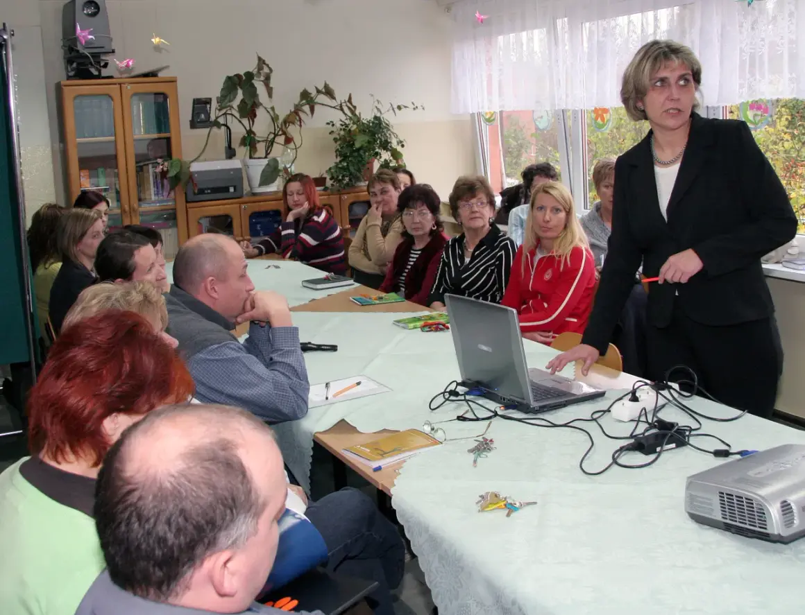 Nauczyciele zebrani na radzie pedagogicznej, dyskutują przy stole z laptopem i projektorem.