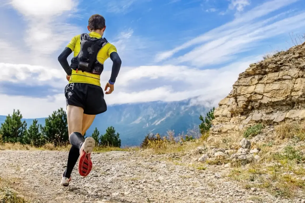 Un coureur en tenue de trail, sac sur le dos, s'élance sur un sentier rocailleux. Le paysage montagneux et le ciel bleu promettent une aventure ultra.