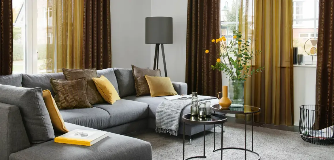 Gemütliches Wohnzimmer mit grauem Sofa, Kissen in Braun und Gelb, sowie moderne Fenstergestaltung mit zweifarbigen Vorhängen.