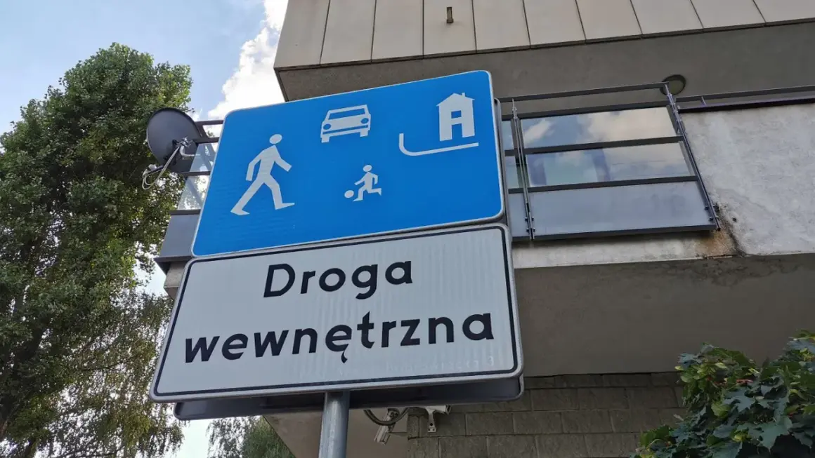 Znak "strefa zamieszkania" informuje o ograniczeniu prędkości. Widoczni piesi, dziecko grające w piłkę, samochód i dom.