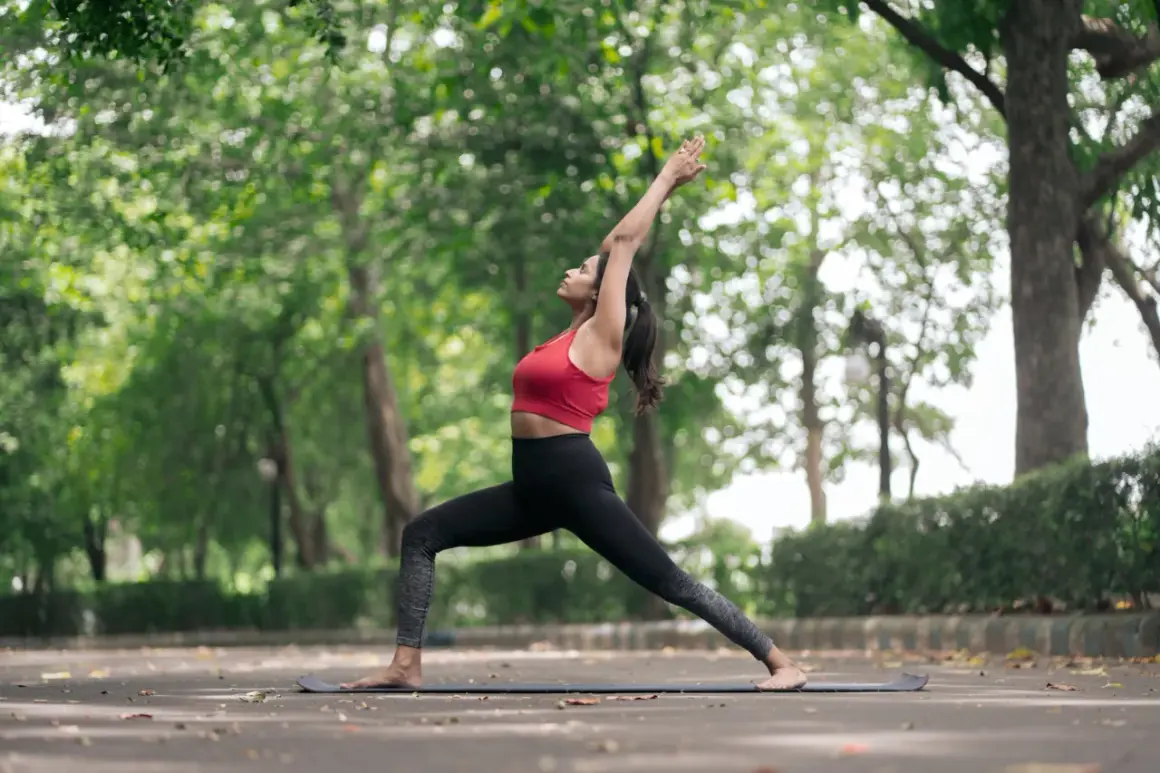 Frau in Krieger-II-Pose, eine von vielen Yoga-Posen, übt im Freien auf einer Matte.