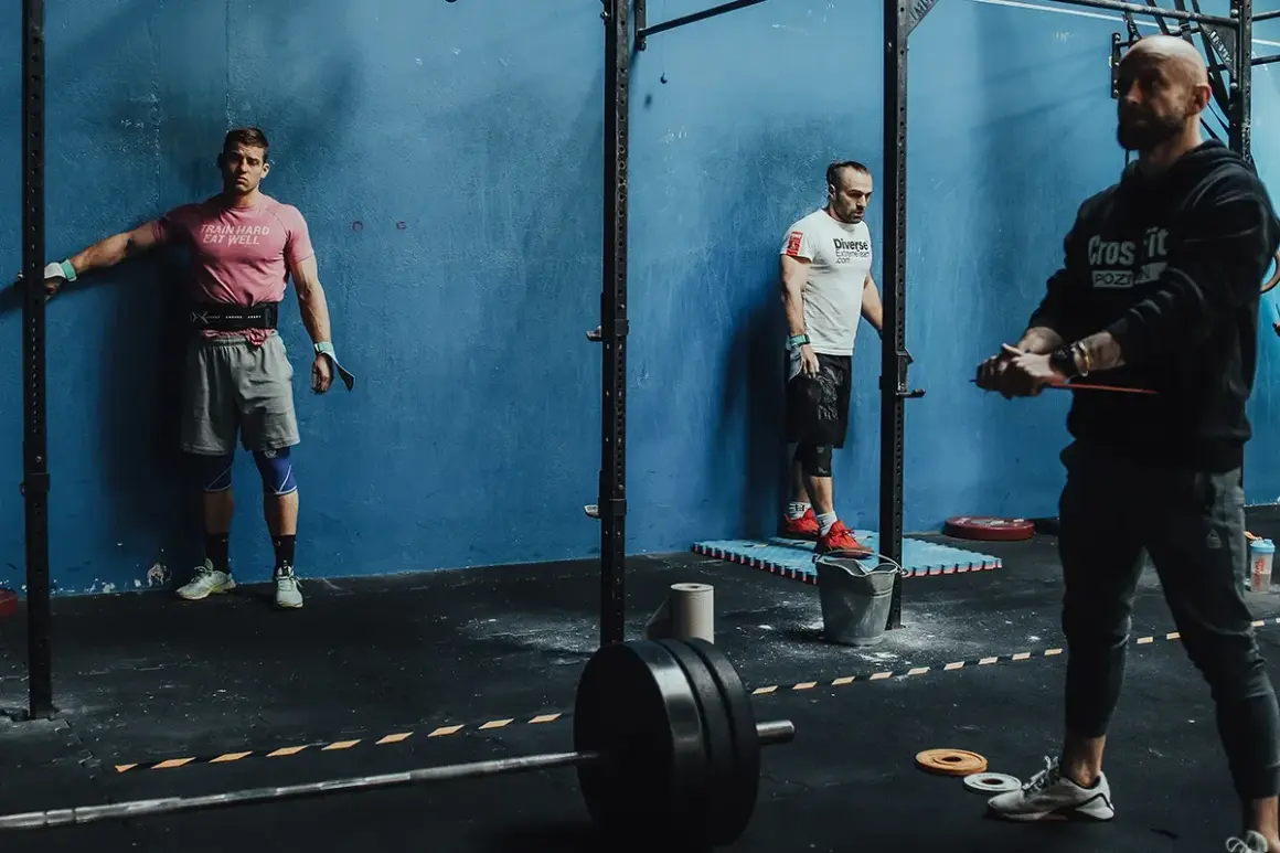 Trzech mężczyzn w siłowni CrossFit, jeden oparty o ścianę, drugi przy stojaku, trzeci z gumą treningową.