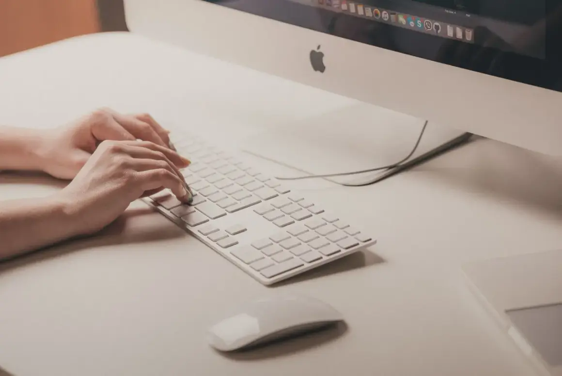 Mani che digitano su tastiera bianca, vicino a un mouse. Attenzione ai sintomi di tendinite da mouse per evitare dolori.