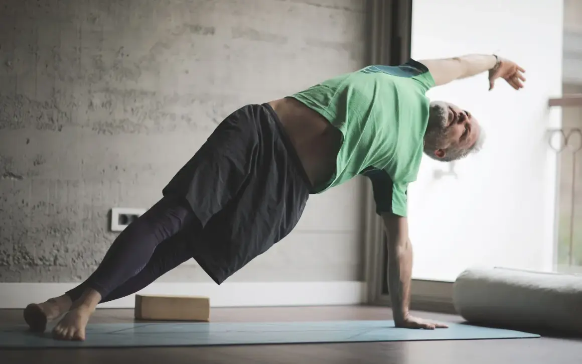 Hombre mayor con barba gris practica yoga para hombres en una esterilla, en postura de plancha lateral.