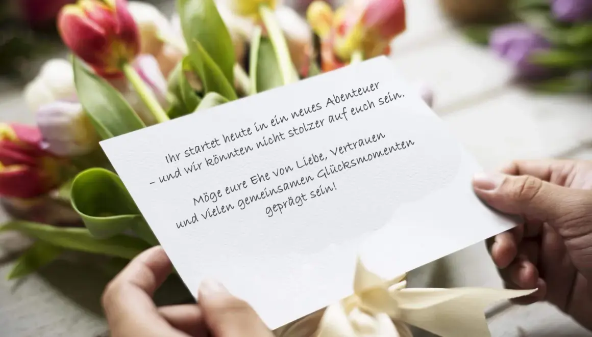 Hände halten eine Karte mit Hochzeitsglückwünschen vor Tulpen. "Ihr startet heute in ein neues Abenteuer – und wir könnten nicht stolzer auf euch sein. Möge eure Ehe von Liebe, Vertrauen und vielen gemeinsamen Glücksmomenten geprägt sein!