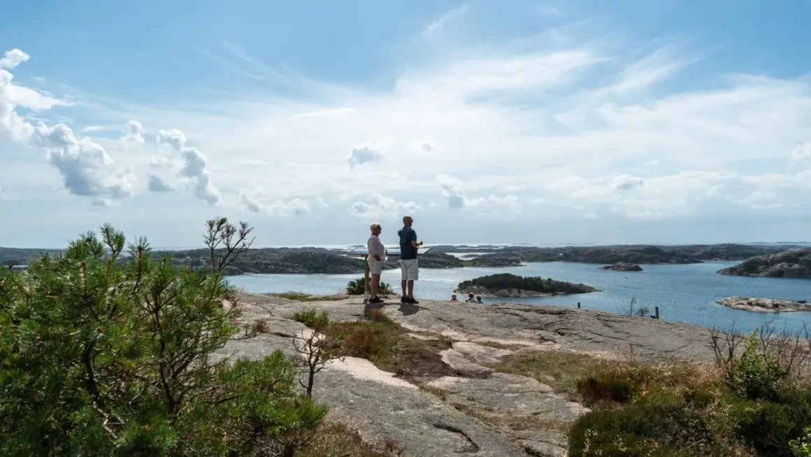 Två personer njuter av utsikten från Vetteberget i Fjällbacka. Vackert väder med moln och sol över skärgården.