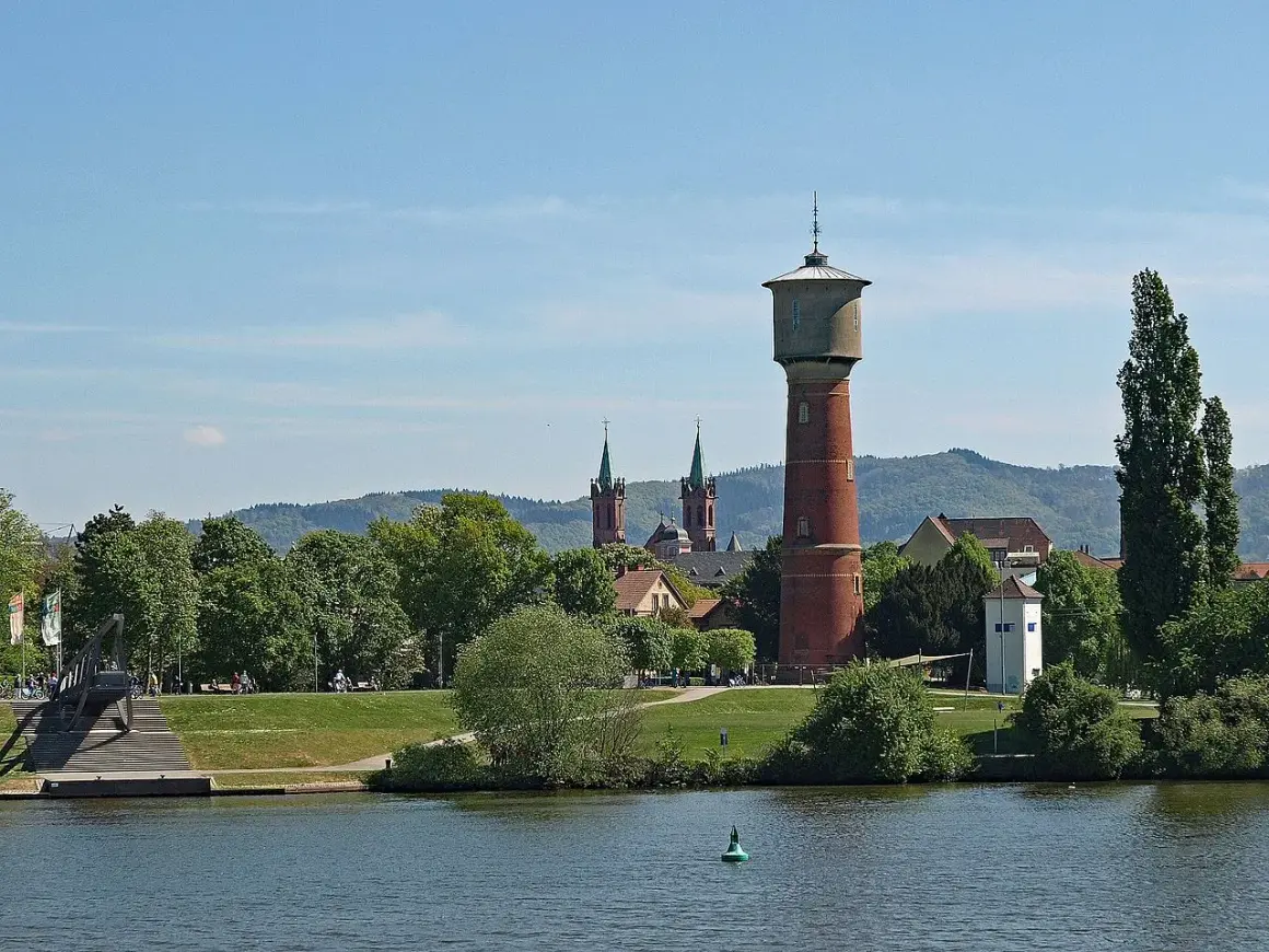 Wasserturm und Kirchtürme über grünen Bäumen, die die ladenburg altstadt säumen, mit Fluss im Vordergrund.
