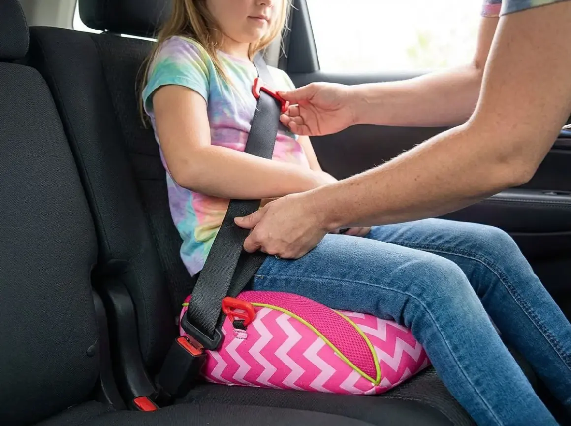 Ein Kind sitzt auf einem pinken Sitzkissen mit Zickzackmuster. Ein Erwachsener befestigt den Sicherheitsgurt, um den Kindersitz zu sichern.