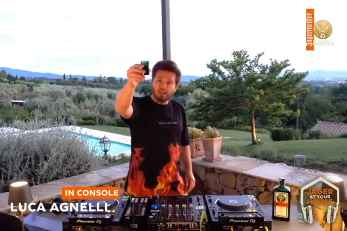 Luca Agnelli, uno dei dj techno famosi, brinda con un bicchierino davanti alla console, con una piscina e un paesaggio collinare sullo sfondo.