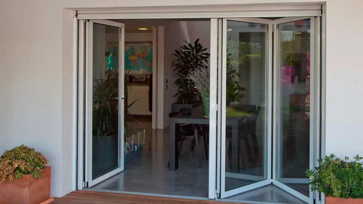 Puertas de cristales plegables que abren a un comedor interior con plantas y una mesa.