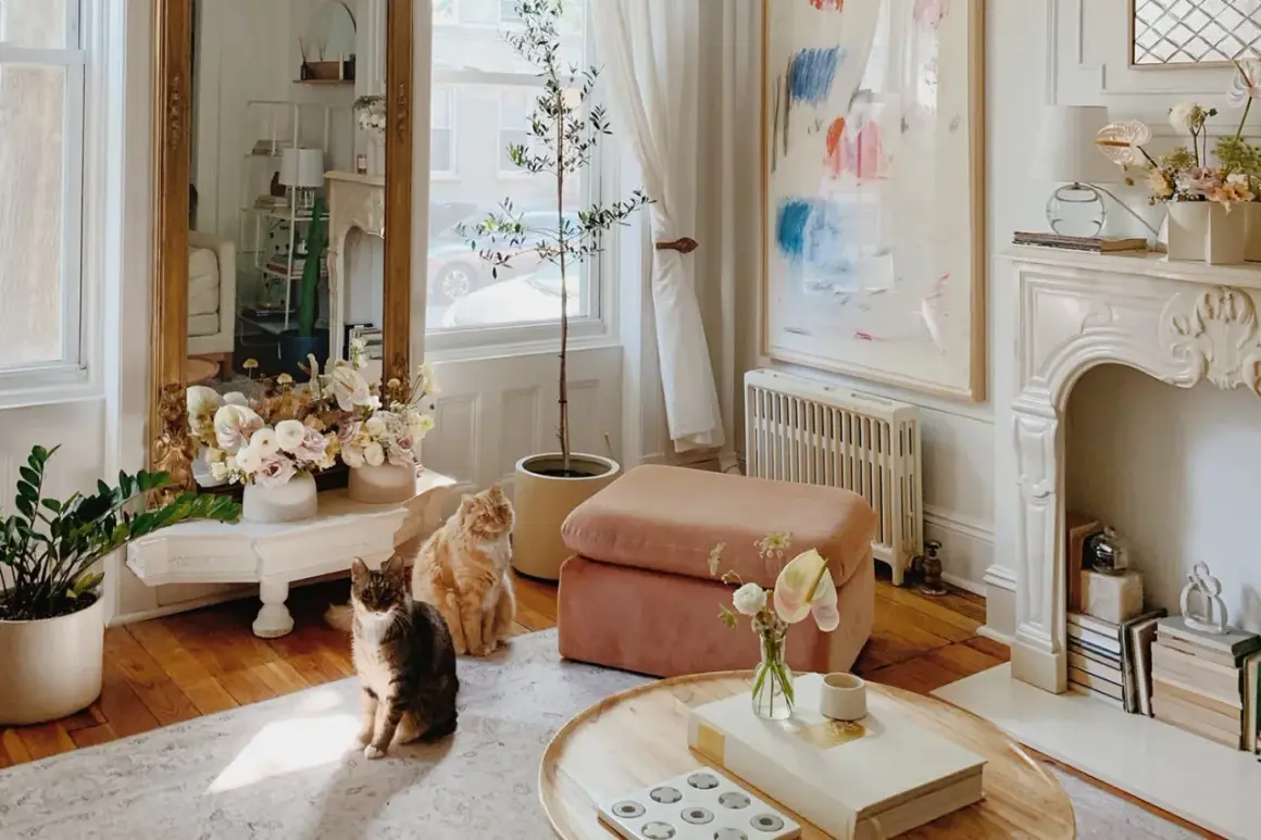 Two cats sit near a tall pier mirror reflecting a bright room with flowers and a painting.