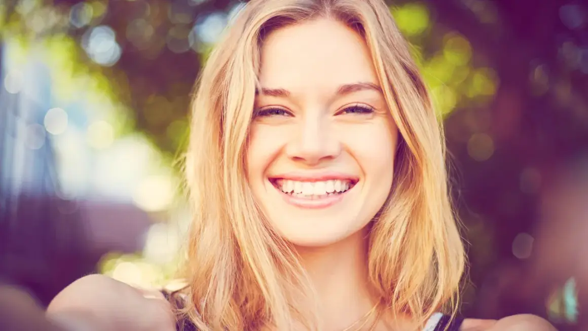 Strahlendes Lächeln einer jungen Frau mit blonden Haaren, die eine natürliche Frisur trägt.