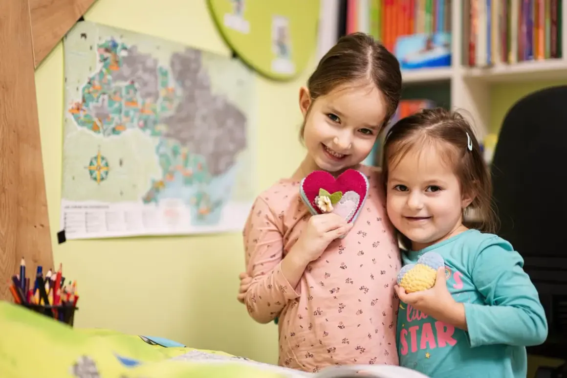 Dwie uśmiechnięte dziewczynki trzymają serduszka. Starsza w różowej bluzce, młodsza w turkusowej. W tle mapa i półka z książkami. Ciekawi ich, ile godzin w zerówce spędzają na zabawie.