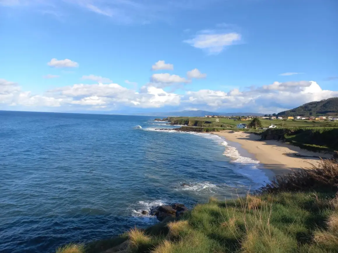 Mar azul y cielo nublado sobre la playa de Xuncos, con casas dispersas en la costa verde.