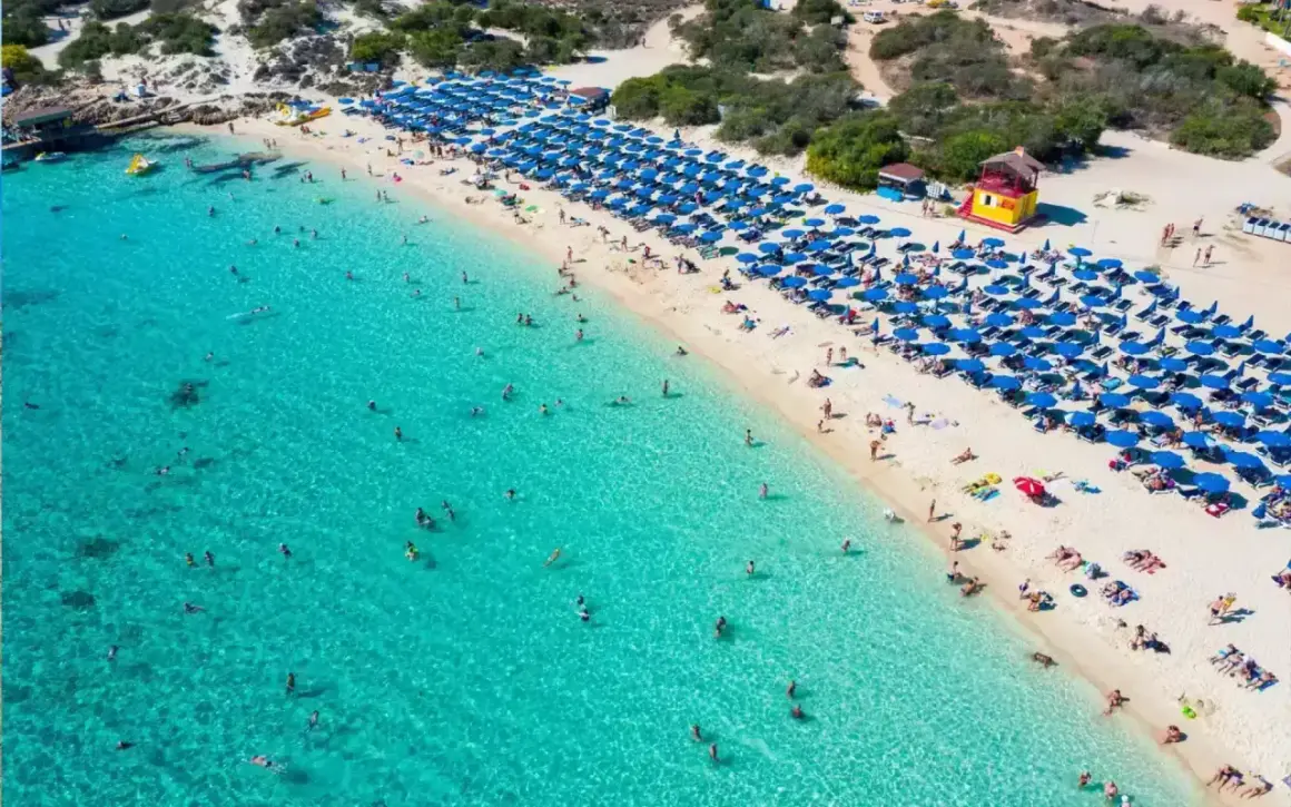 Słoneczny dzień na plaży Makronissos. Turkusowa woda, biały piasek i mnóstwo niebieskich parasoli. Ludzie kąpią się i opalają.
