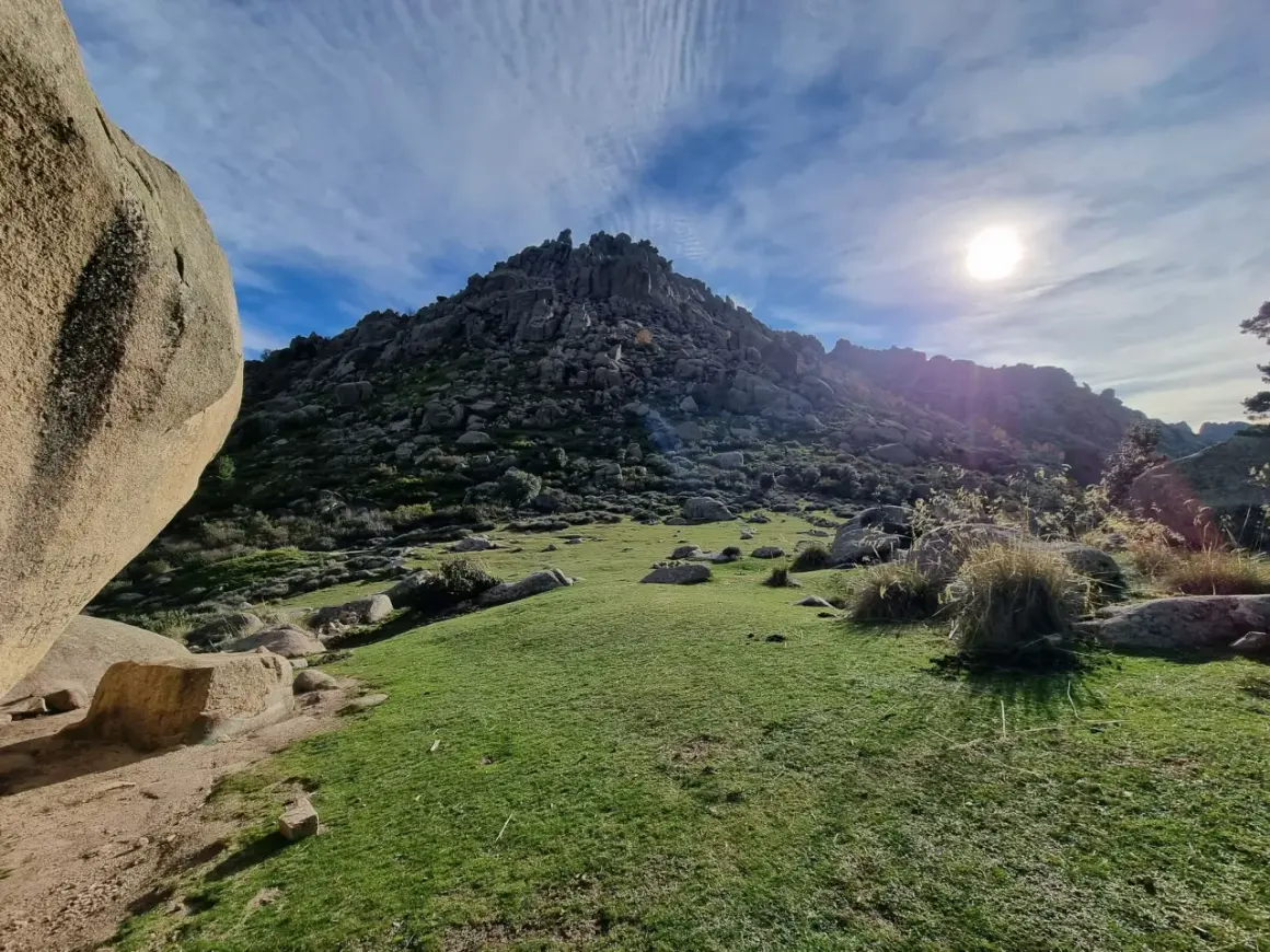 Majestuosa montaña rocosa en la ruta Soto del Real, con un prado verde y el sol brillando en un cielo azul con nubes.