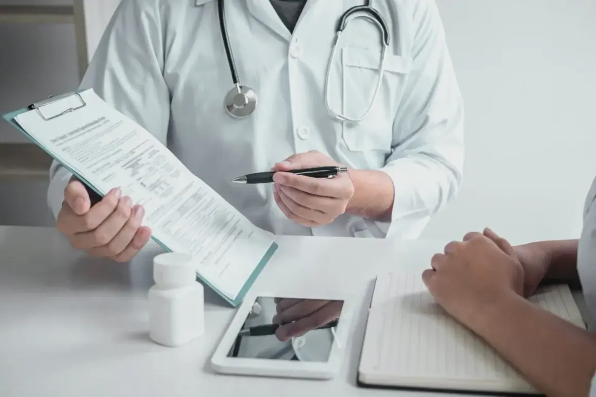 Lekarz w białym kitlu omawia z pacjentem dokumenty, prawdopodobnie dotyczące zwolnienia lekarskiego w Norwegii. Na stole leżą tablet i notatnik.