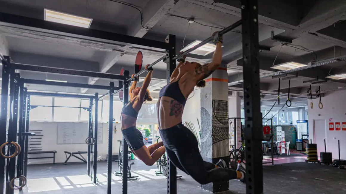 Dwie kobiety wykonują podciąganie na drążku w siłowni. To część treningu, który może być elementem "fran crossfit co to jest".