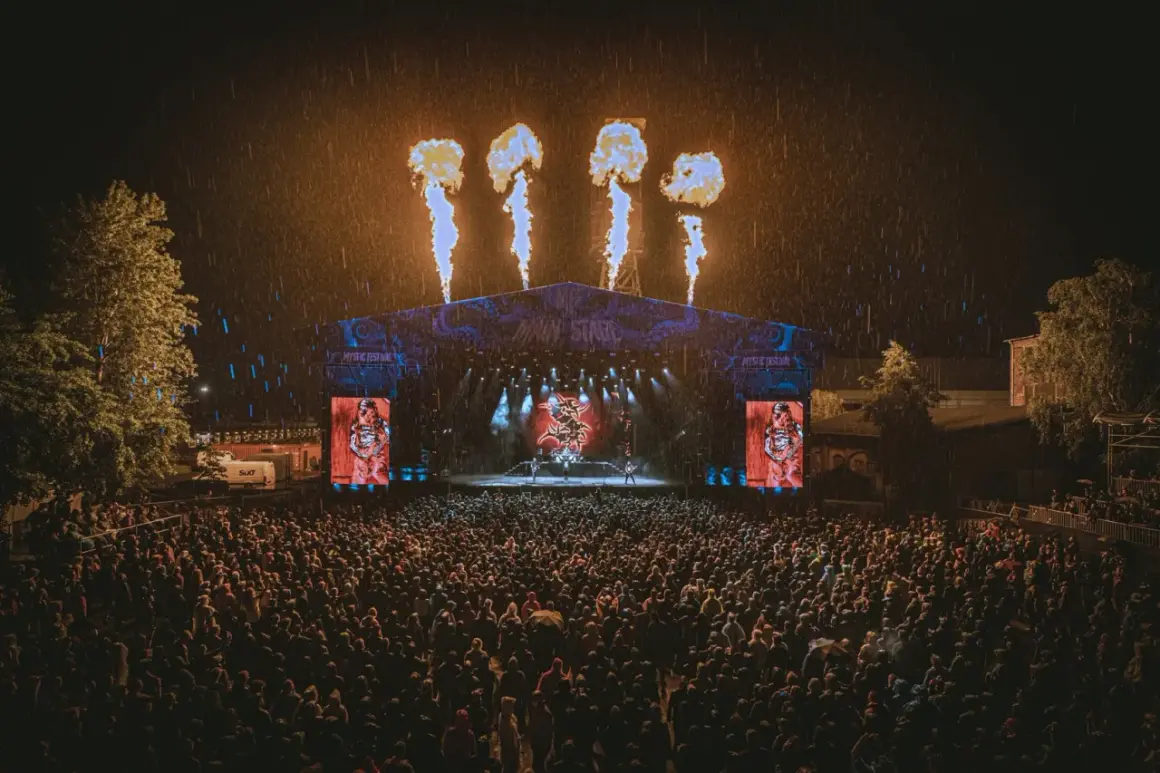 Koncert na Mystic Festival w deszczu, z płomieniami nad sceną i tłumem fanów.