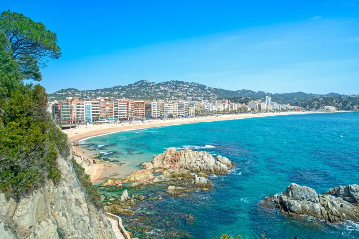 Widok na plażę i zatokę w Lloret de Mar. W oddali widać zabudowania, a na pierwszym planie skaliste wybrzeże i turkusowe morze. Idealne miejsce na wakacje, niedaleko lotniska.