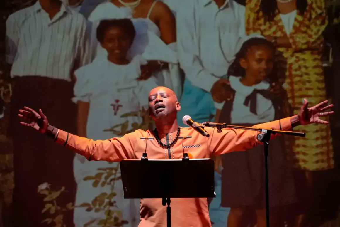 A man in an orange shirt sings with his arms outstretched in front of a projection of a family.