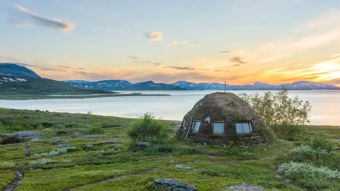 En grästorvstuga med kors på toppen vid en sjö i Padjelanta nationalpark, Sverige, under en solnedgång.