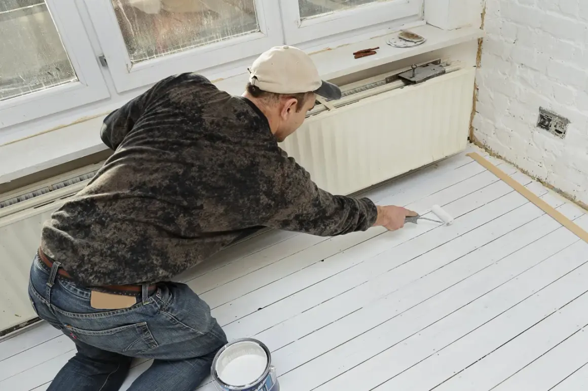 Un homme peint un plancher en bois blanc. Il utilise un rouleau et un pot de peinture. La question "peut on peindre du stratifié" est pertinente pour ce type de projet.