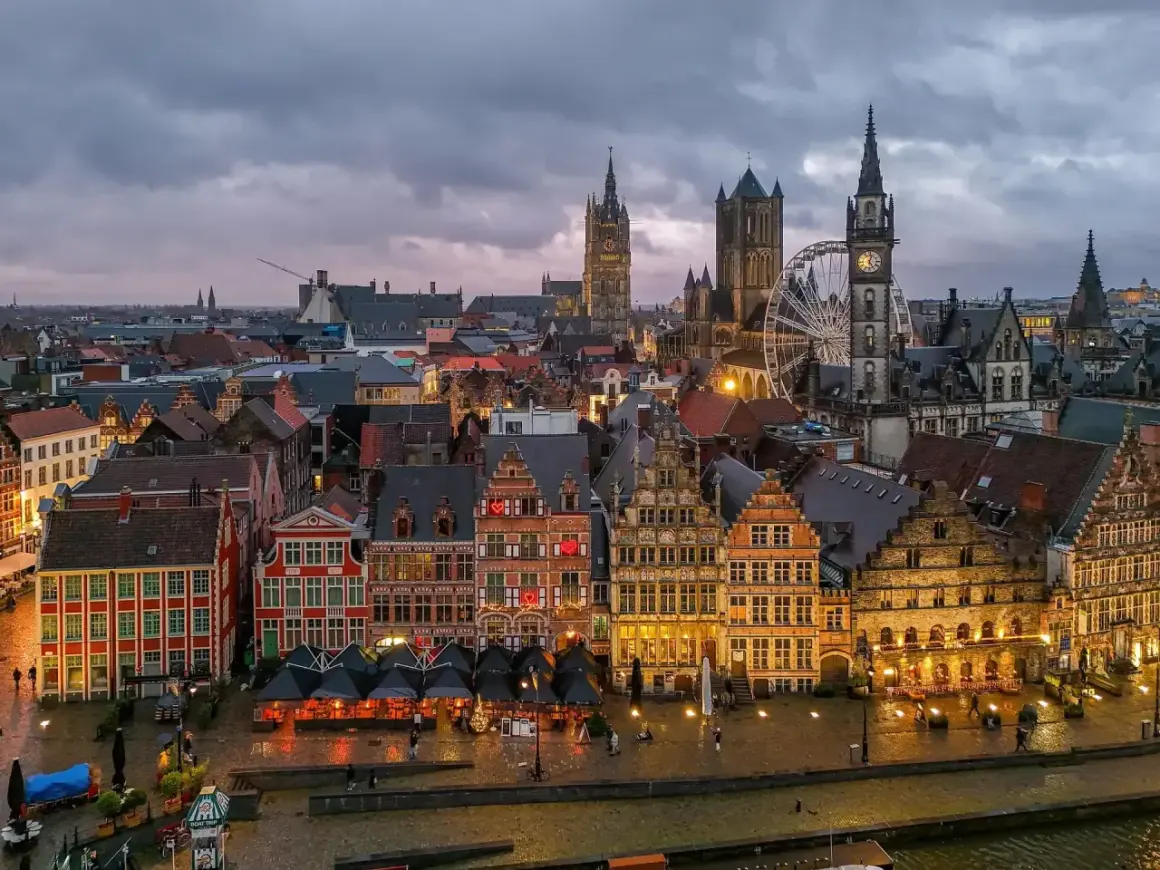 Gryning över Gent, Belgien. Historiska byggnader längs en kanal, med ett pariserhjul och klocktorn som reser sig mot en molnig himmel.