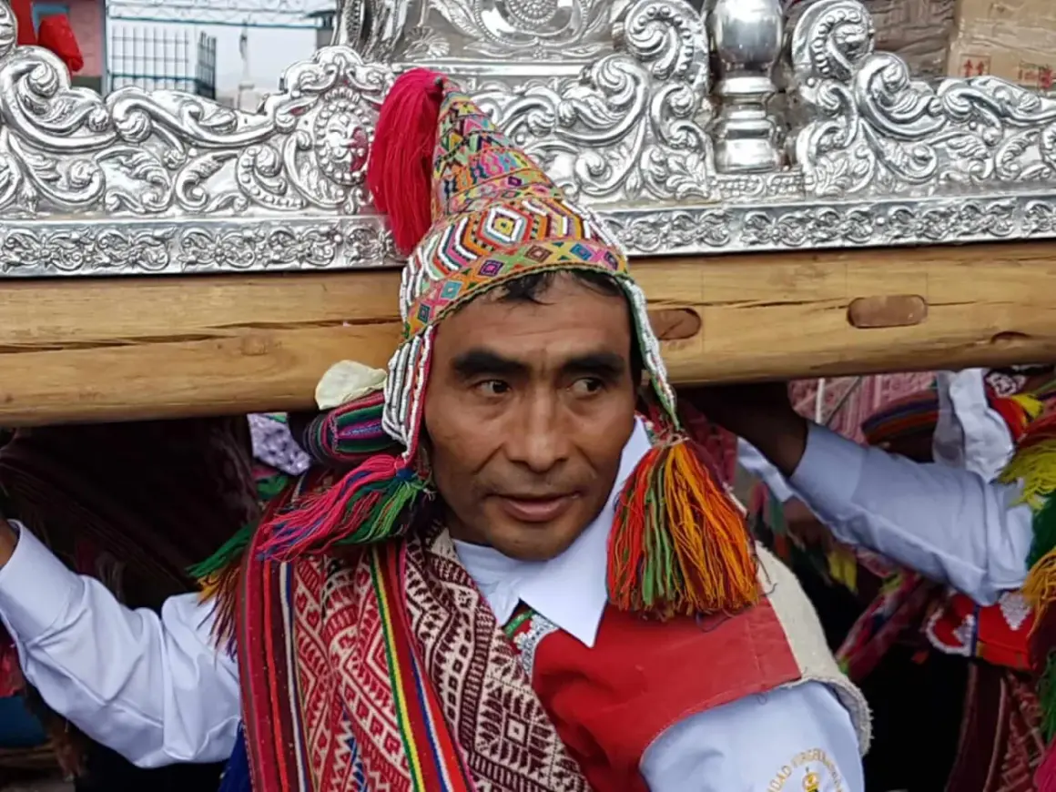 Kolorowy strój i tradycyjna czapka zdobią jednego z mężczyzn niosących ozdobną ramę. To **Indianie z Cuzco** podczas uroczystości.