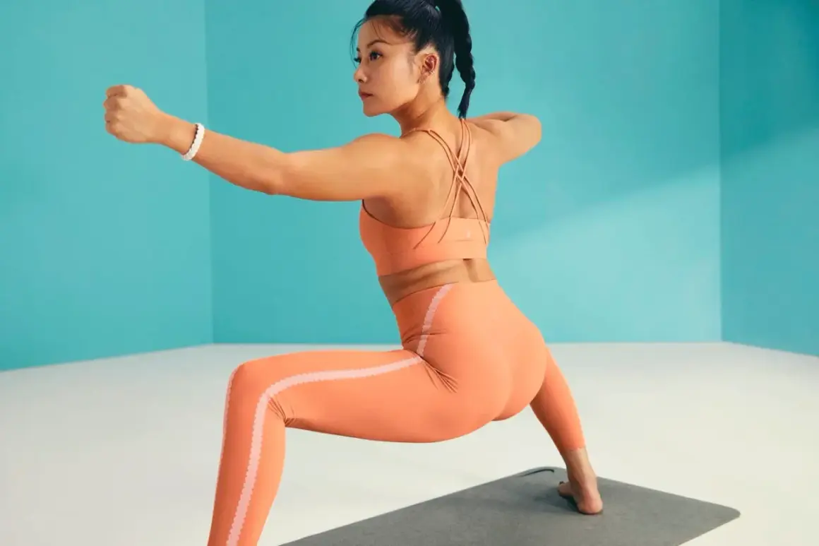 Mujer en ropa deportiva naranja practicando asanas de pie, con un brazo extendido y el otro doblado.