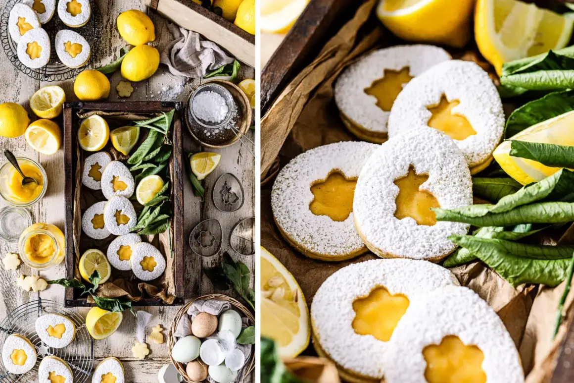 Zuckrige Kekse mit leuchtend gelbem Lemon Curd gefüllt, umgeben von frischen Zitronen und grünen Blättern.