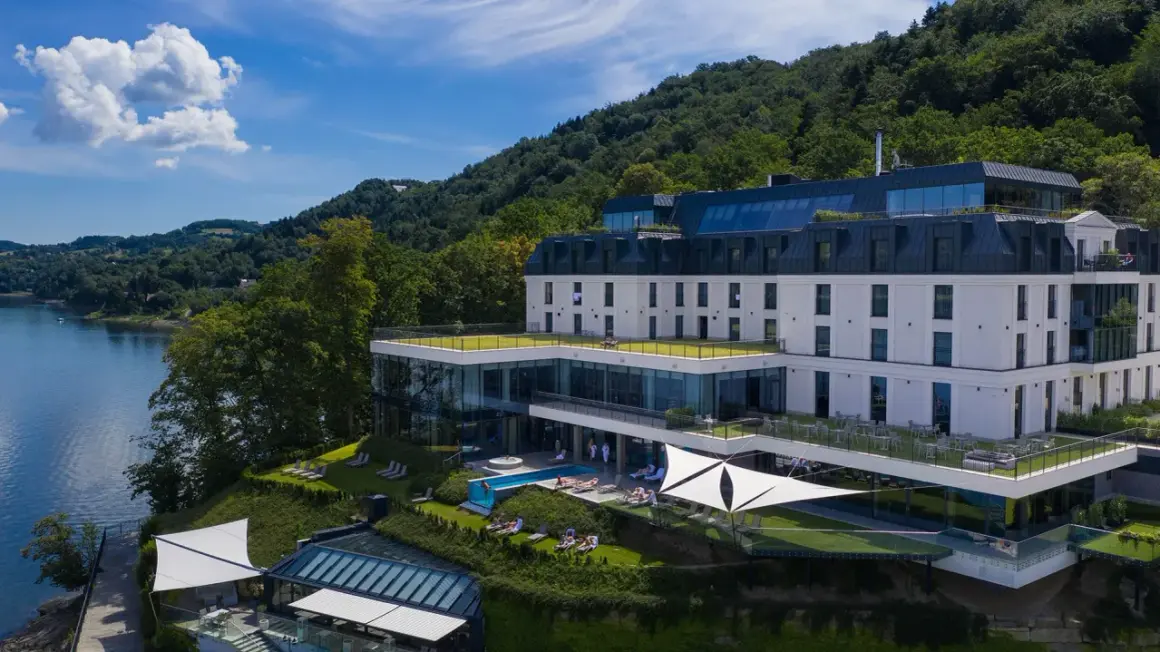 Nowoczesny hotel nad jeziorem Rożnowskim, z basenem i tarasami, otoczony zielenią.
