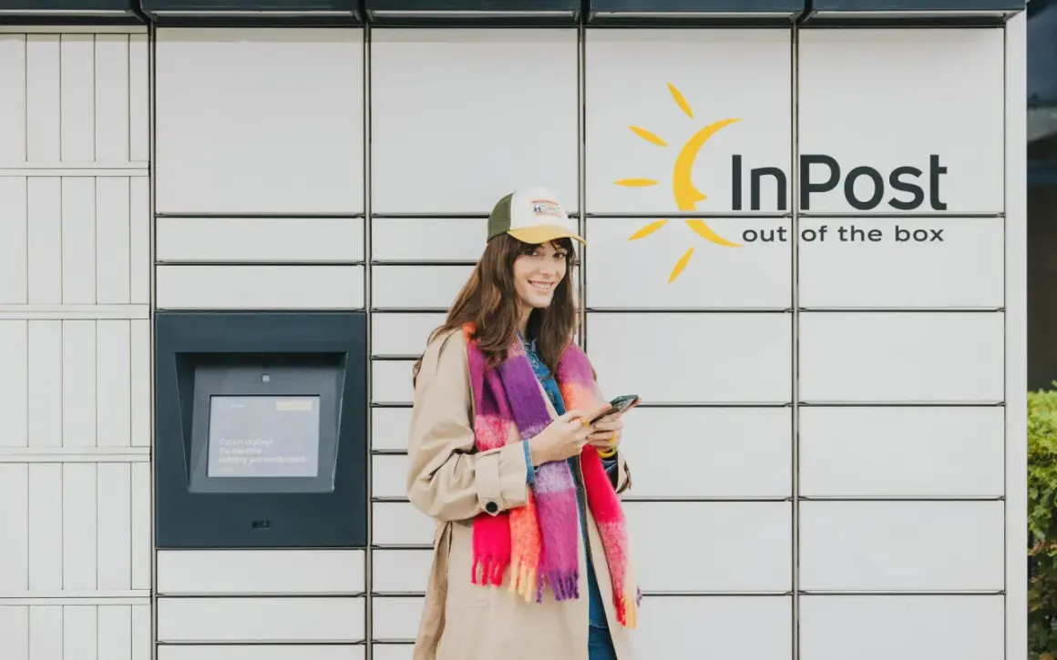 Uśmiechnięta kobieta z telefonem stoi przy paczkomacie InPost. Wygodne akcje InPost to jej codzienność.