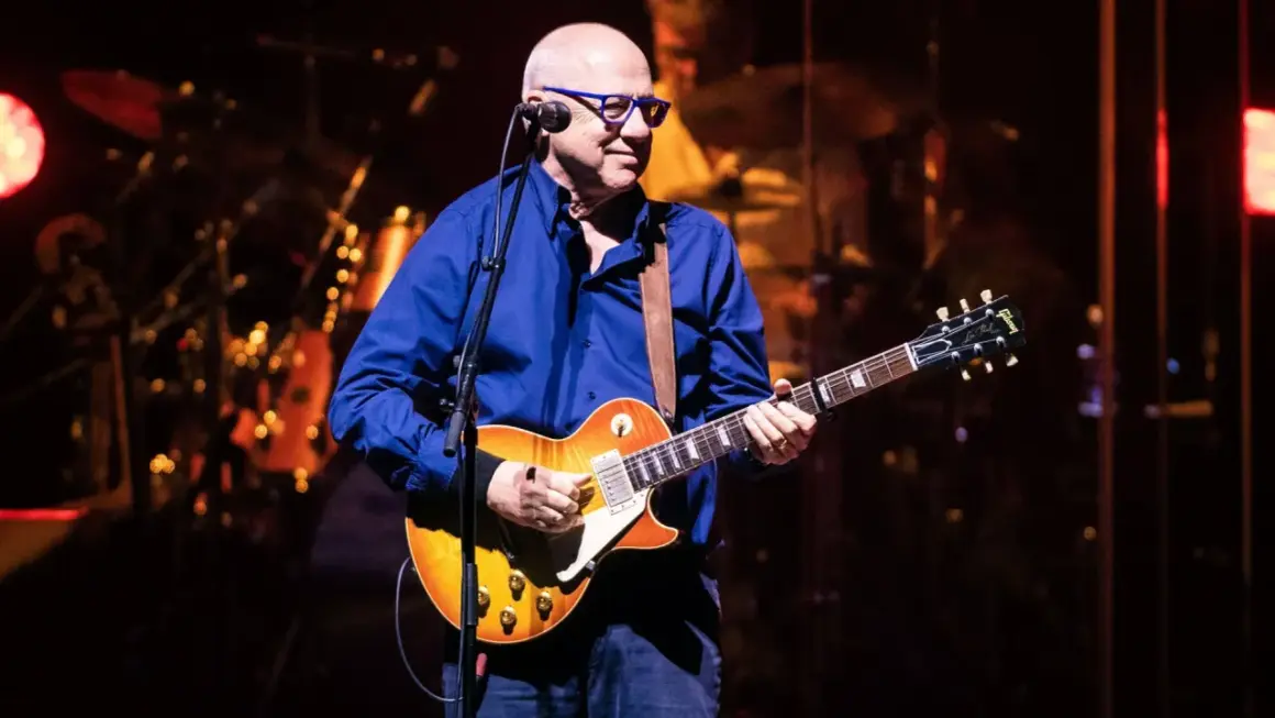 Mark Knopfler, w okularach i niebieskiej koszuli, gra na gitarze Gibson. Jego muzyka, jak na jego albumy, brzmi w tej chwili na żywo.