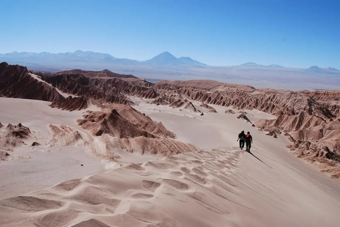 Dos excursionistas caminan por una duna en el desierto de Atacama, con formaciones rocosas y montañas al fondo bajo un cielo azul.