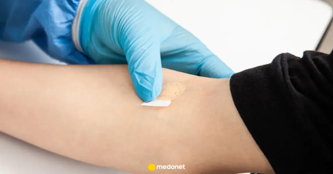Ręka w niebieskiej rękawiczce zakłada plaster na ramię. Może to być związane z badaniem poziomu hormonów stresu.