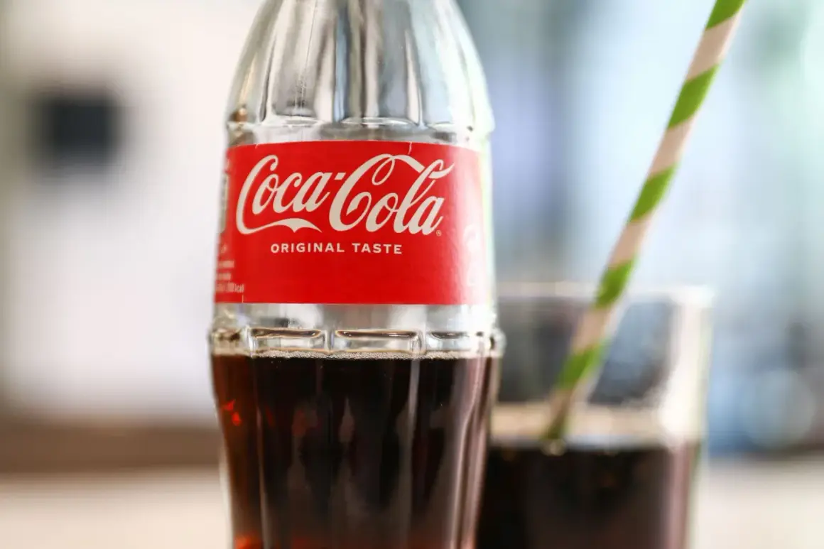 Szklana butelka Coca-Coli z czerwono-białą etykietą i słomką w tle.