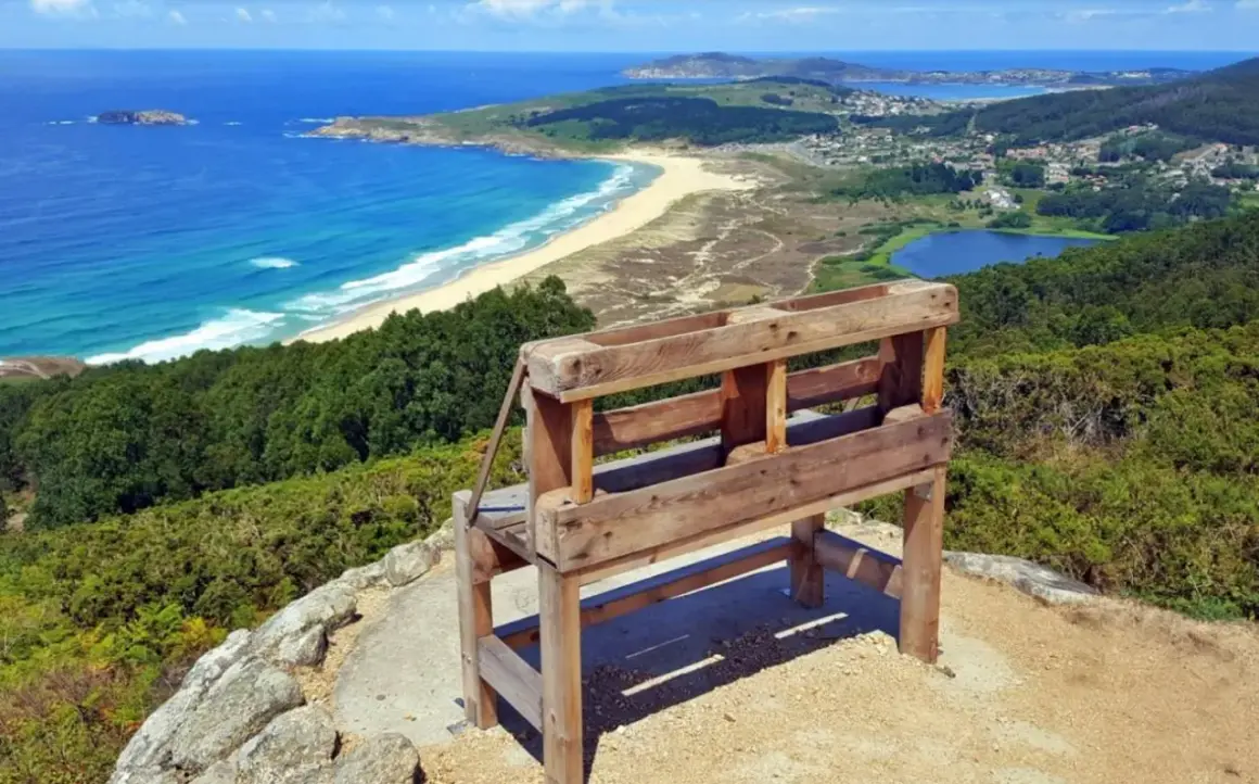 Banco de madera en un monte ventoso con vistas a una playa, el mar azul y un lago.