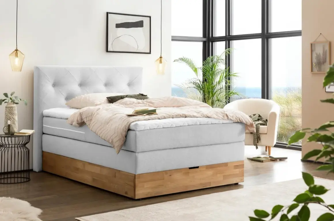 Ein hellgraues Boxspringbett mit Holzsockel und einem gesteppten Kopfteil. Die Nachteile eines Boxspringbetts, wie z.B. die Höhe, sind hier nicht ersichtlich.