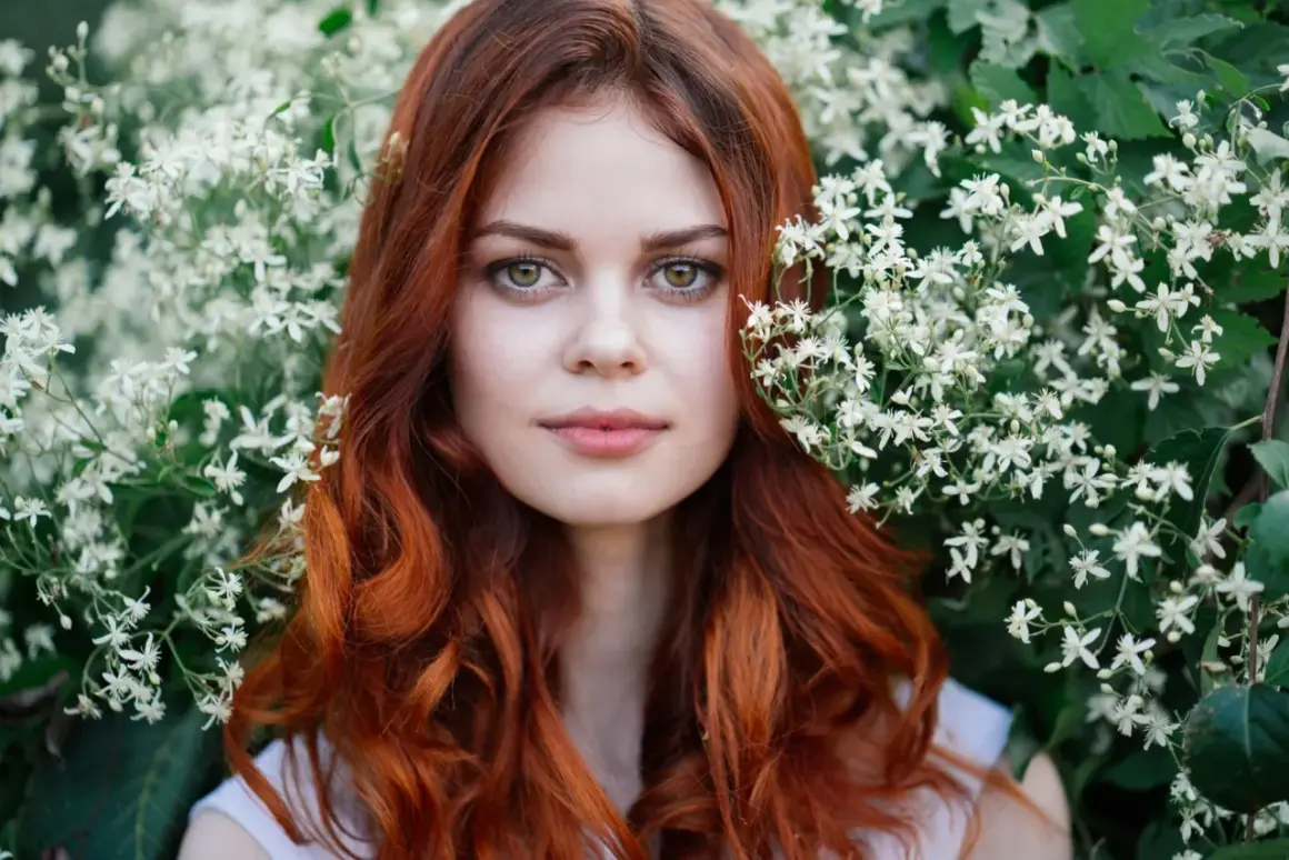 Eine Frau mit leuchtend roten, bunten Haaren blickt aus einem Meer von weißen Blüten.