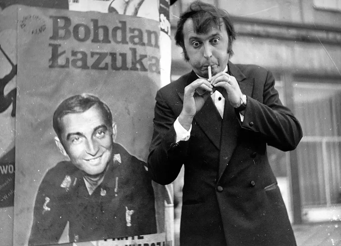 Bohdan Łazuka w garniturze, z papierosem w ustach, obok plakatu z jego podobizną.