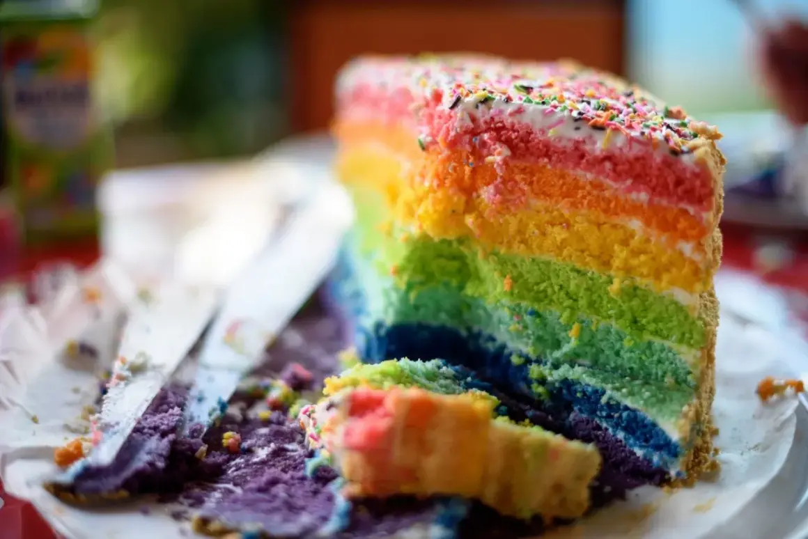 Ein Stück bunter **Regenbogen Kuchen** mit bunten Streuseln. Ein Stück ist bereits angeschnitten und liegt daneben.