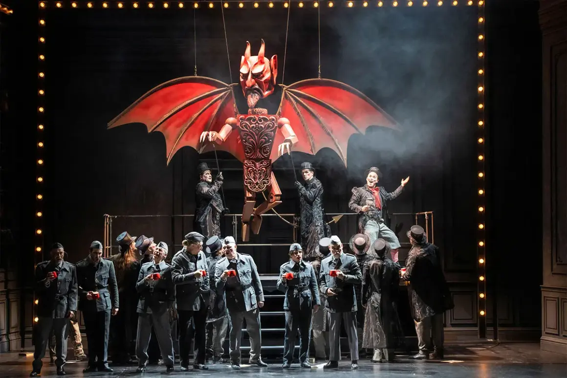 Bühnenszene der Oper Wuppertal: Eine Teufelsmarionette schwebt über Darstellern in Uniformen und Zylindern.