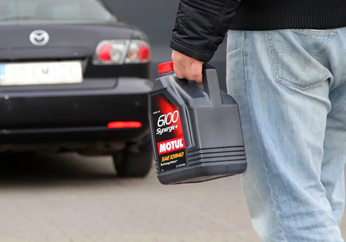 Mężczyzna niesie kanister z olejem silnikowym Motul 6100 Synergie+ SAE 10W40. Za nim stoi czarny samochód.