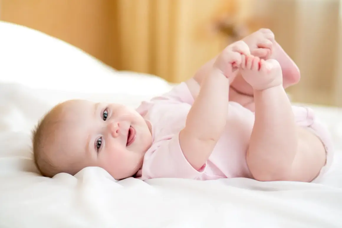 Ein fröhliches Baby von 8 Wochen liegt auf dem Rücken und spielt mit seinen Füßen.
