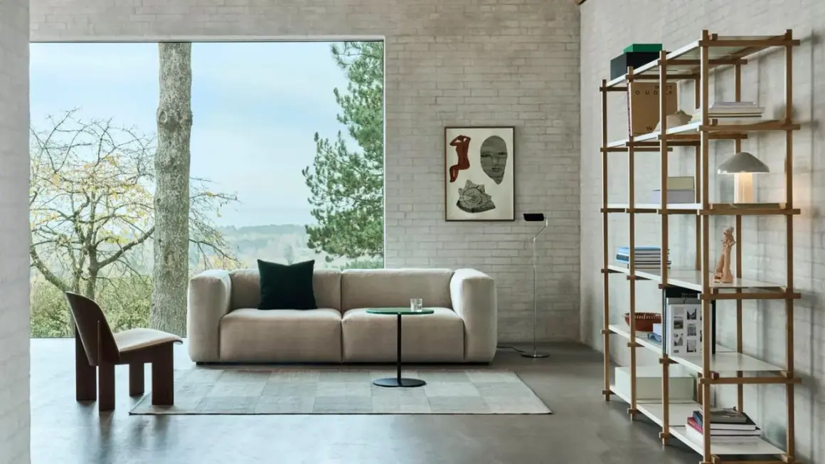 Gemütliches Wohnzimmer mit beigem Sofa, einem Holzsessel und einem Regal voller Bücher. Der Stil ist modern, mit einem Hauch von millennial grey in den Ziegelwänden.