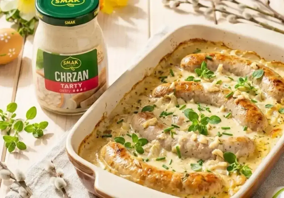 Biała kiełbasa w sosie chrzanowym, zapiekana w naczyniu, z dodatkiem ziół. Obok słoik sosu chrzanowego.
