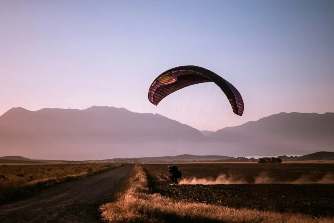 Parapentista aterrizando en un campo polvoriento al atardecer, rodeado de montañas. Una escena de turismo activo.